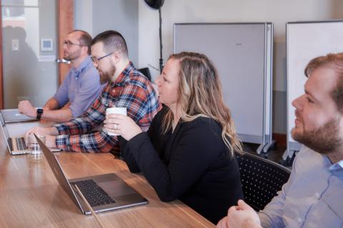 Lisa sits in front of her laptop holding a coffee cup, in a row of coworkers all looking toward a presentation