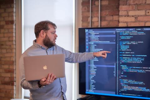 Sam stands and points at code on presentation monitor while holding laptop