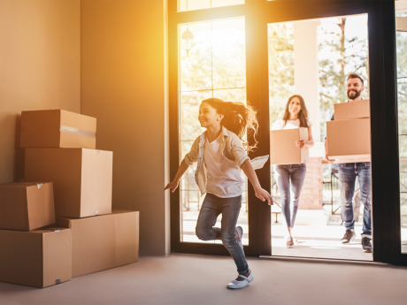 Girl runs happily into new home filled with boxes, followed by parents carrying boxes